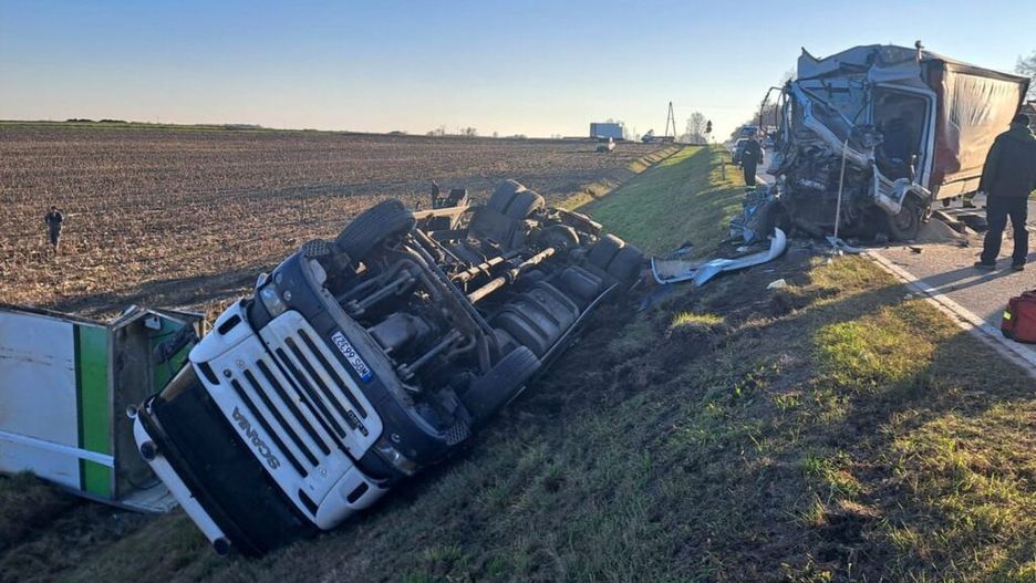 Czarny ciężarówka leży na boku w rowie, obok uszkodzona naczepa, a w tle widoczni ratownicy na wiejskiej drodze.