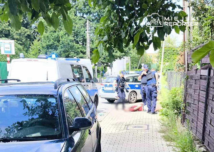 Policjanci udzielają pomocy leżącej osobie na chodniku obok zaparkowanych samochodów w miejskim otoczeniu.