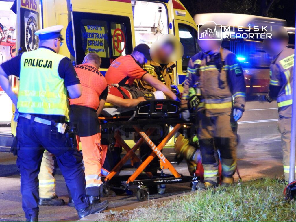Grupa ratowników medycznych i strażaków transportuje poszkodowanego na noszach obok karetki na drodze nocą.