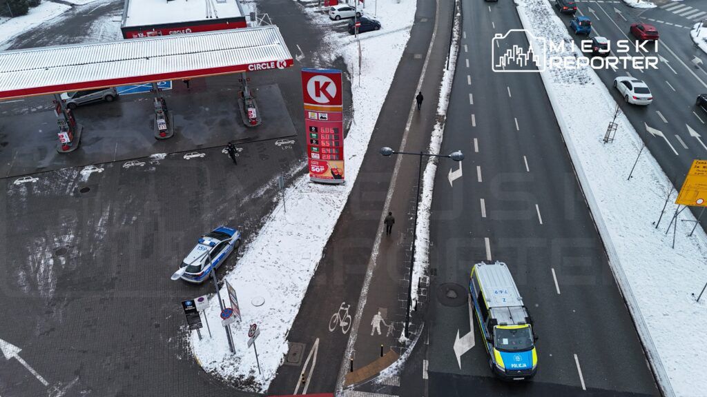 Policyjny radiowóz zaparkowany obok stacji benzynowej w zimowym otoczeniu, obok przechodnia idącego chodnikiem.