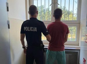 Policjant w mundurze stoi za mężczyzną w koszulce, obaj patrzą przez okno w pomieszczeniu z kratami.