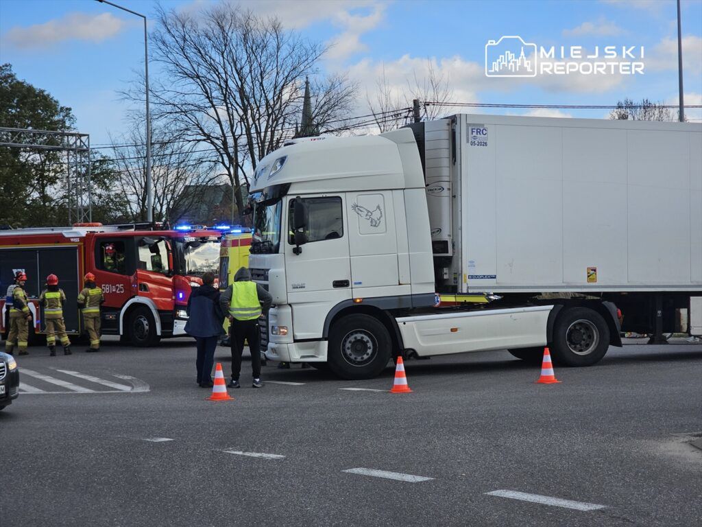 Czterech strażaków w mundurach rozmawia z mężczyzną w kamizelce odblaskowej obok ciężarówki, przy włączonym wozie strażackim.