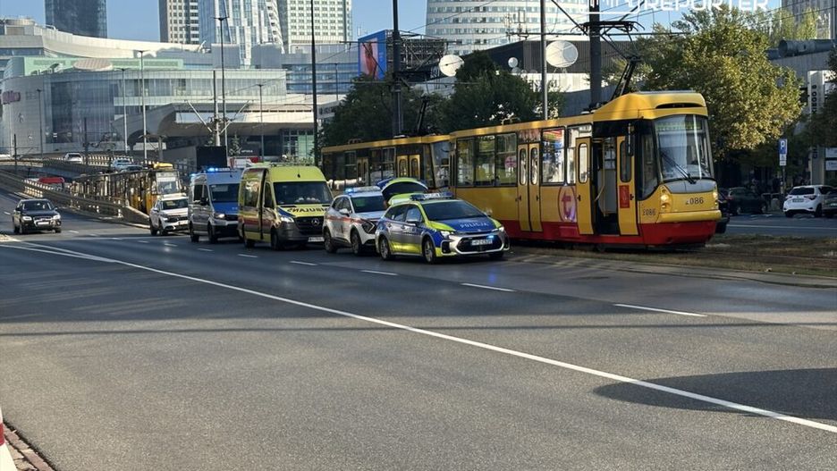 Wielu funkcjonariuszy policji oraz karetka blokują ruch na ulicy obok tramwaju w nowoczesnym otoczeniu miejskim.