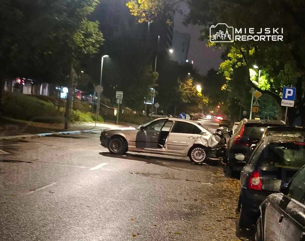 Zniszczony samochód osobowy stoi na ulicy, otoczony innymi zaparkowanymi pojazdami, w nocy.