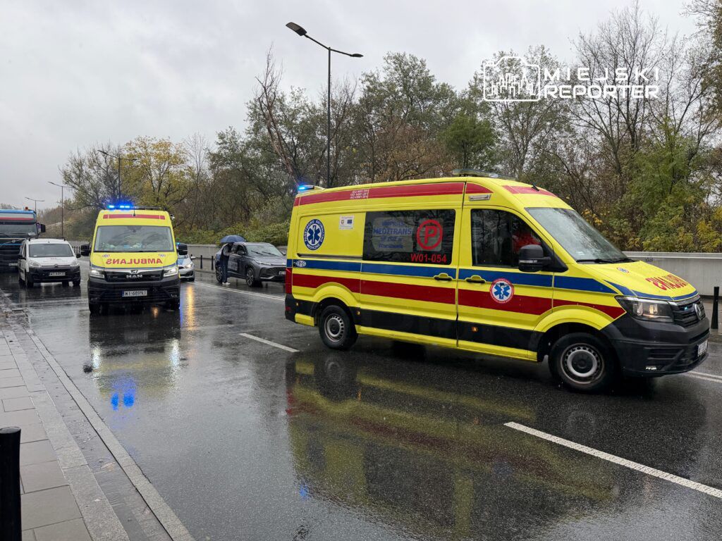 Ambulans z włączonymi sygnałami świetlnymi zatrzymany na deszczowej ulicy, obok innych pojazdów.
