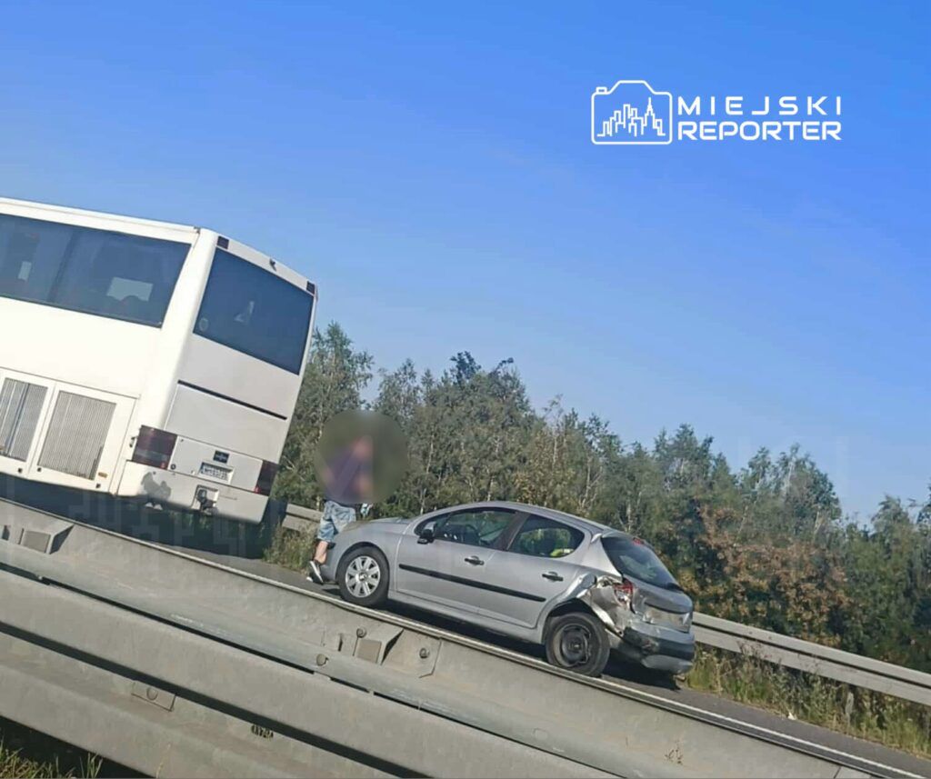 Mężczyzna w szortach stoi obok uszkodzonego samochodu na poboczu drogi, obok przejeżdżającego autobusu.