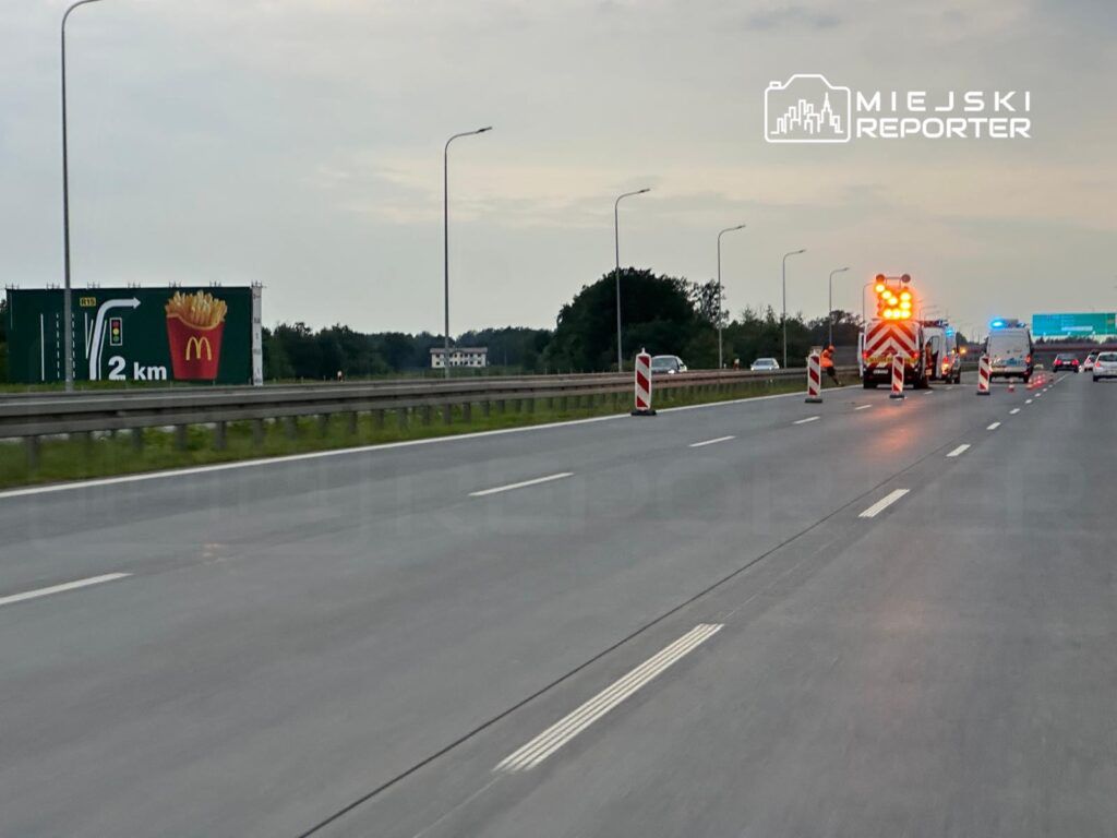 Na drodze widoczna jest akcja służb ratunkowych z włączonymi sygnałami świetlnymi, obok reklama McDonald's informująca o odległości 2 km.
