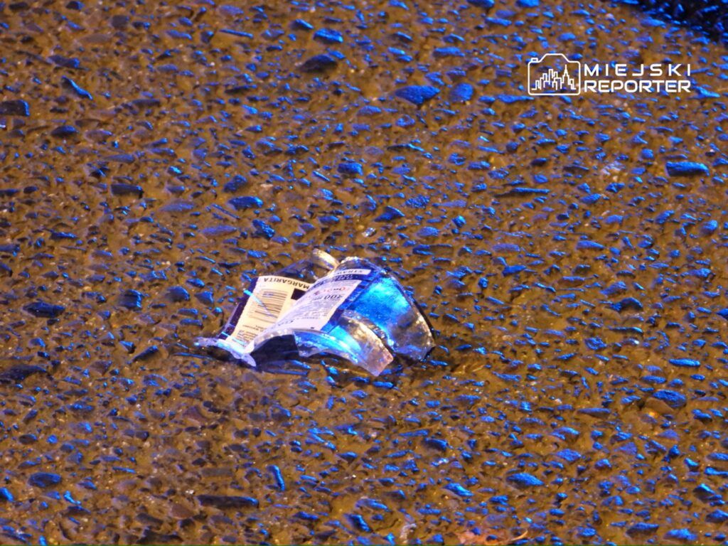 Szkło butelki leży na mokrej nawierzchni drogi, oświetlone niebieskim światłem.