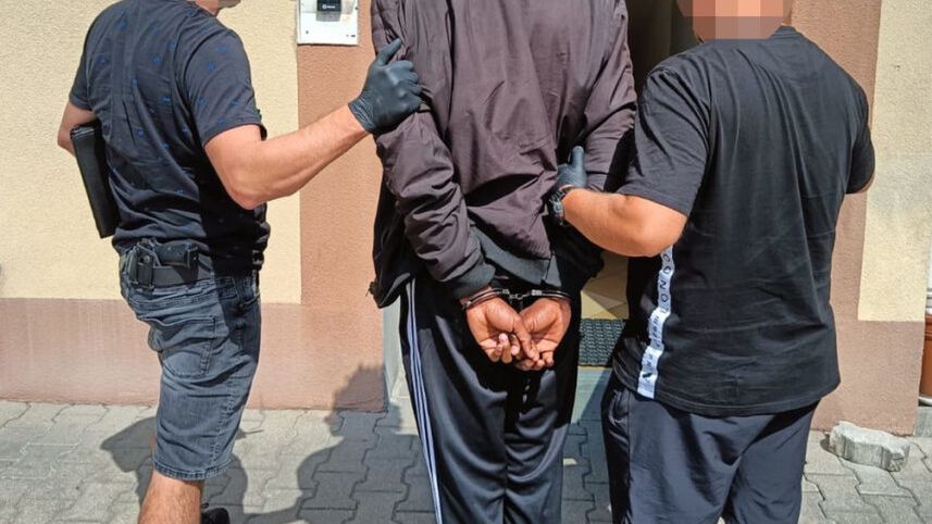 Mężczyzna w dresie prowadzony przez dwóch funkcjonariuszy policji w kierunku budynku.