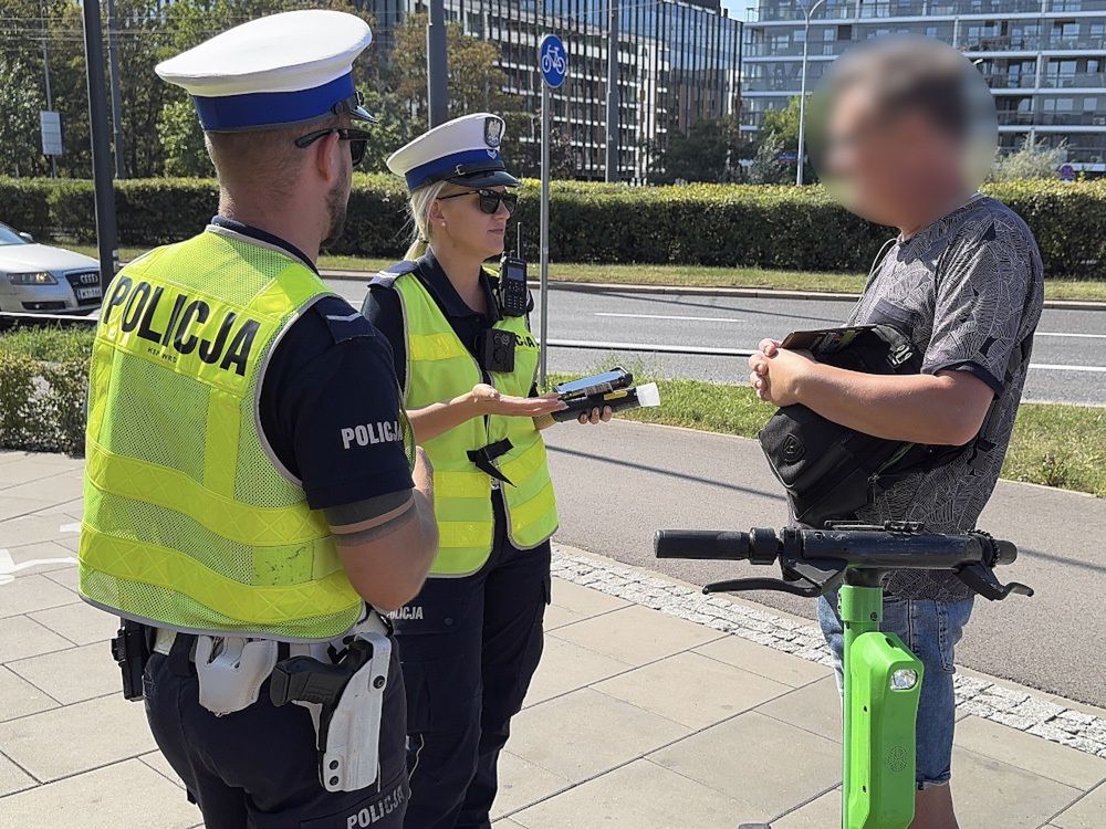 Policjantka w kamizelce odblaskowej rozmawia z mężczyzną stojącym obok zielonego hulajnogi na chodniku.