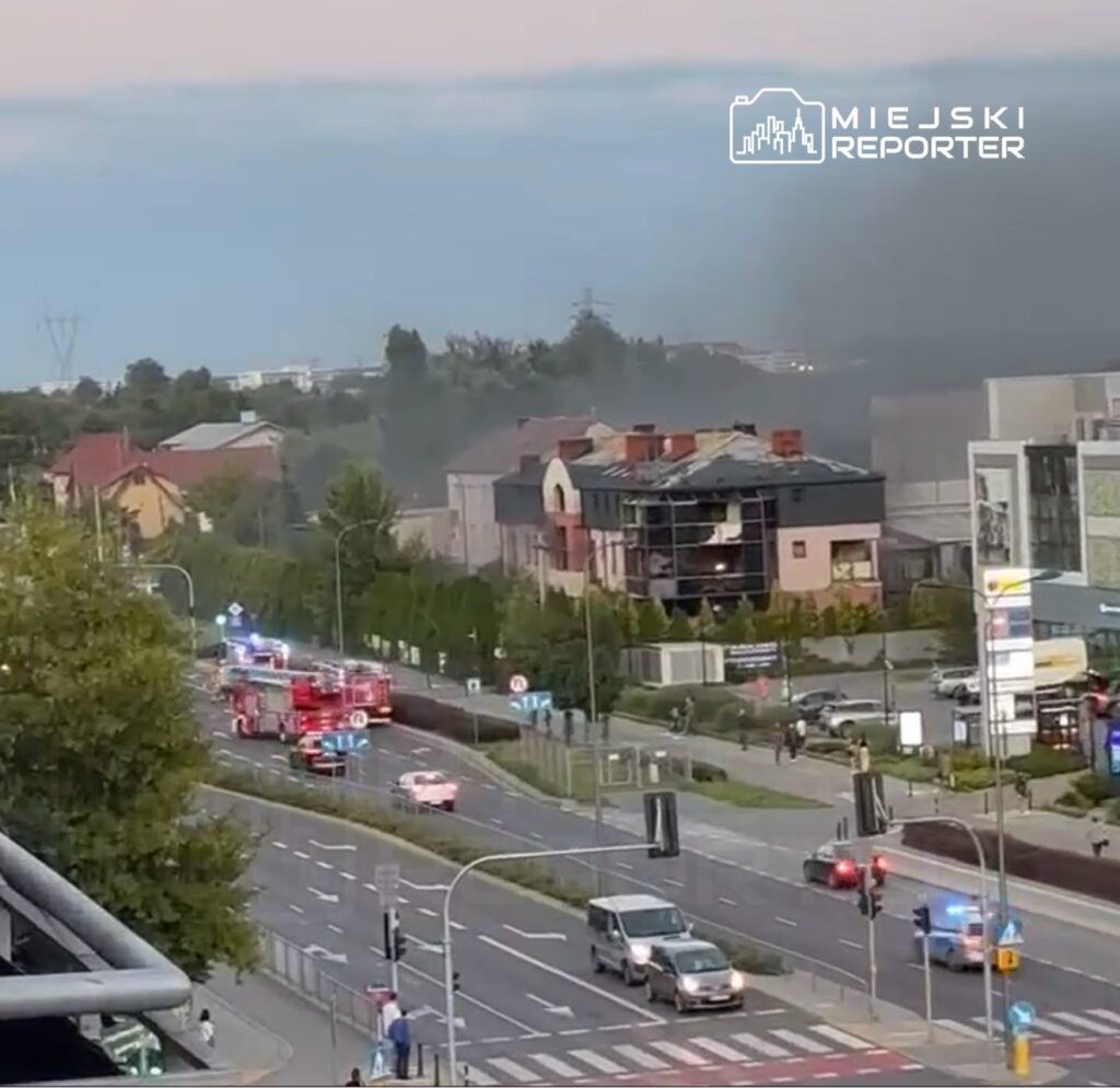 Wielka akcja ratunkowa z wozami strażackimi na ruchliwej ulicy, w tle widoczny dym z płonącego budynku.