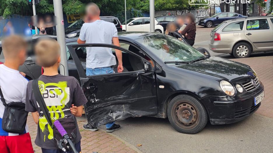 Mężczyzna rozmawia z innym mężczyzną obok uszkodzonego czarnego samochodu na ruchliwej ulicy, obok grupy dzieci.