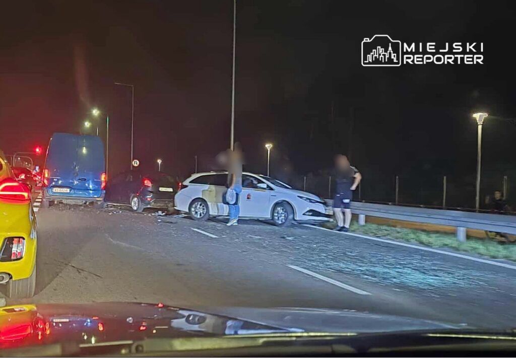 Kolizja drogowa z udziałem kilku pojazdów na nocnej jezdni, z uszkodzonymi samochodami i stojącymi obok osobami.
