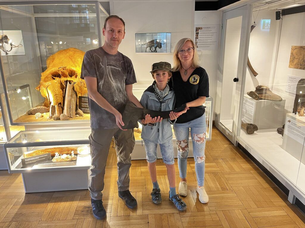 Mężczyzna, chłopiec i kobieta trzymają eksponat w muzeum, w tle widoczne skamieniałości i wystawy.