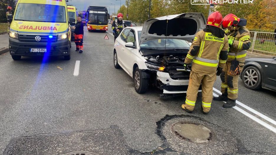 Dwie karetki pogotowia oraz strażacy sprawdzają uszkodzony samochód osobowy na ruchliwej ulicy.
