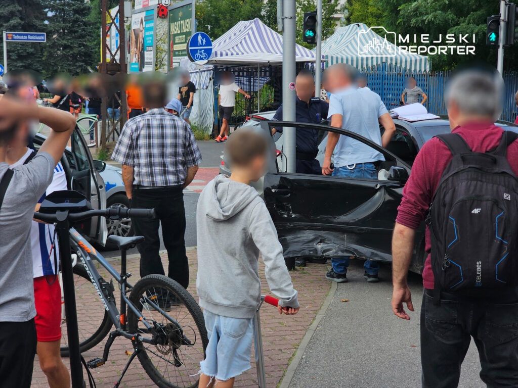 Grupa osób gromadzi się wokół uszkodzonego samochodu na ulicy, obok policyjnego radiowozu.