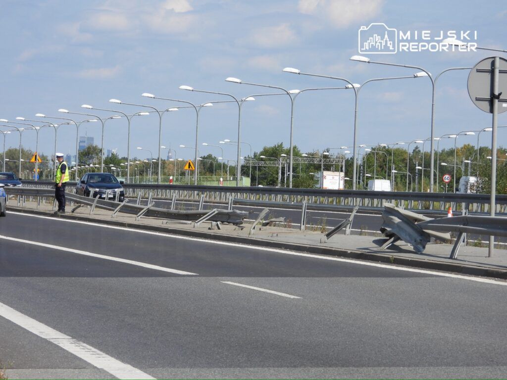 Policjant w kamizelce odblaskowej kieruje ruchem na drodze, obok uszkodzonego bariery ochronnej.