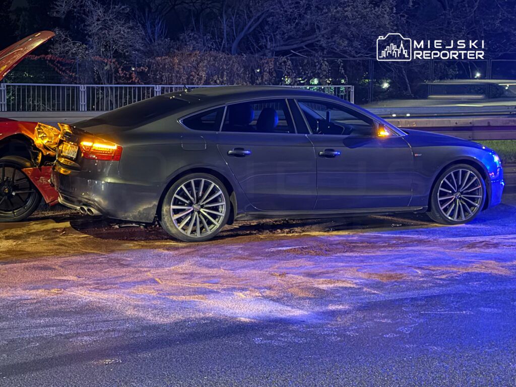 Czarny samochód osobowy zaparkowany obok uszkodzonego pojazdu, w tle widać nocne oświetlenie ulicy.