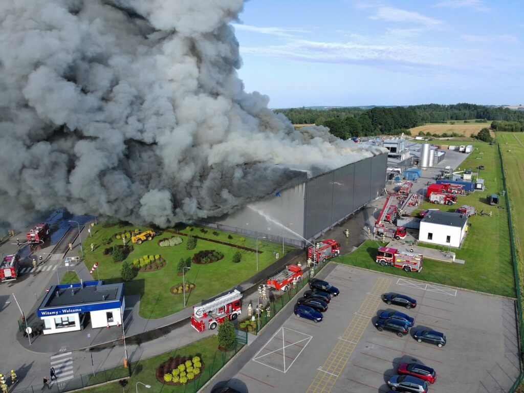 Gęsty dym unoszący się z płonącego magazynu, obok strażacy gaszący ogień, w tle nowoczesny budynek przemysłowy.