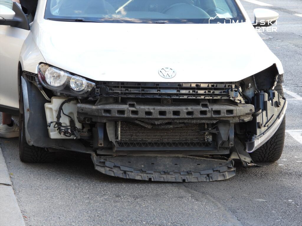 Uszkodzony biały samochód osobowy z brakiem przedniego zderzaka, stojący na parkingu.