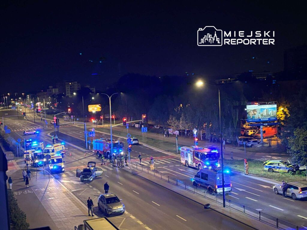 Na drodze nocą odbywa się akcja ratunkowa z udziałem straży pożarnej i karetek, w tle widać sygnalizację świetlną.