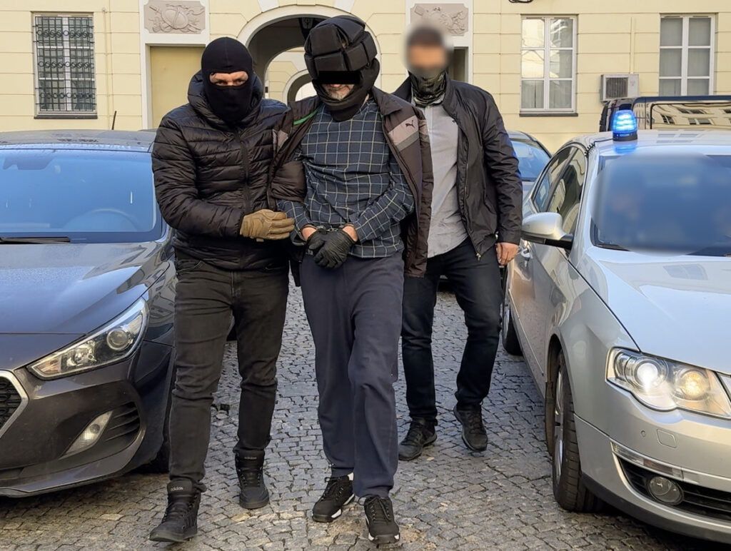 Mężczyzna w kajdankach prowadzony przez dwóch uzbrojonych funkcjonariuszy w pobliżu zaparkowanych samochodów.