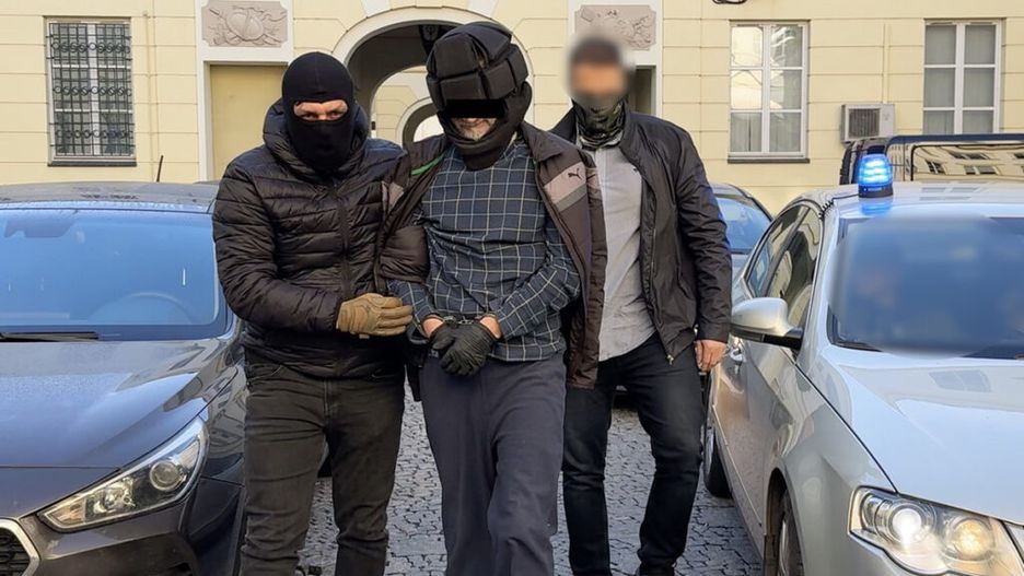 Mężczyzna w kajdankach prowadzony przez dwóch uzbrojonych funkcjonariuszy w pobliżu zaparkowanych samochodów.