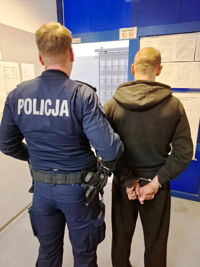 Policjant w mundurze prowadzi zatrzymanego mężczyznę w kajdankach w korytarzu komisariatu.