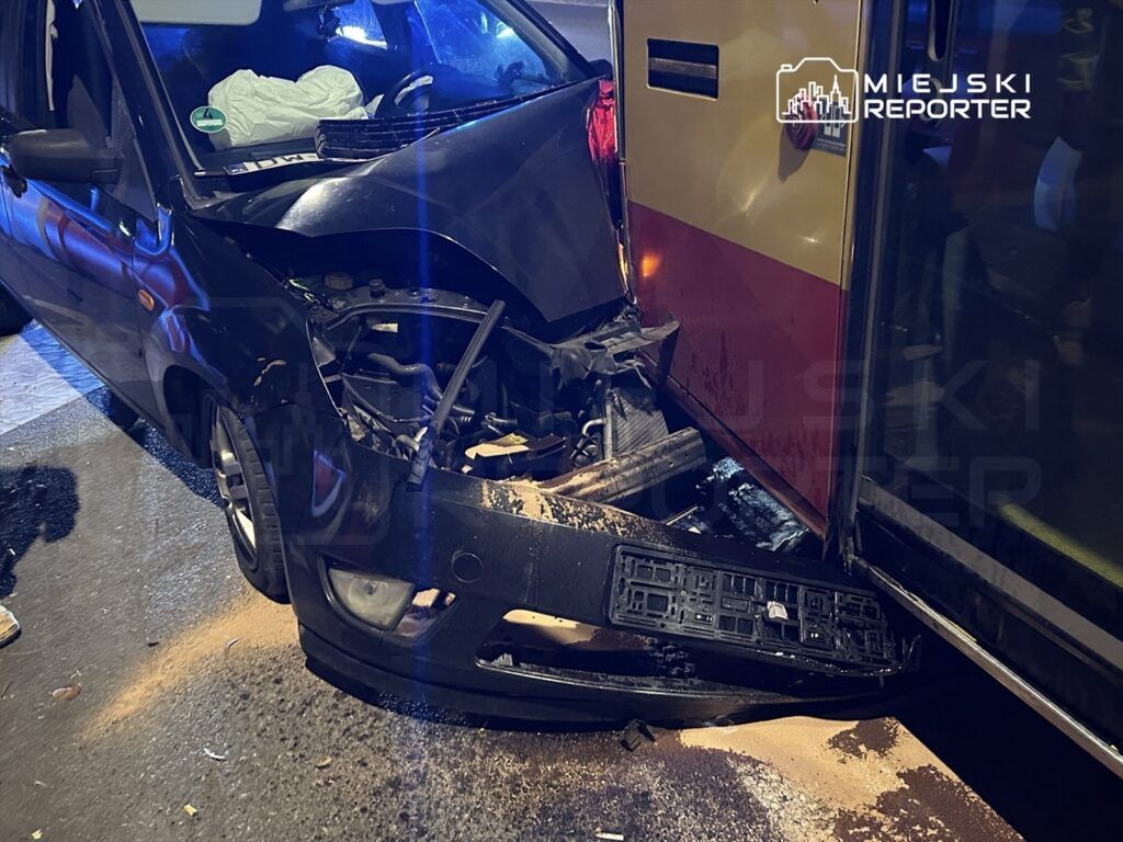 Czarny samochód osobowy z uszkodzoną przednią częścią zderza się z karetką pogotowia na oświetlonej ulicy.