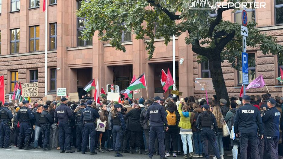 Tłum demonstrantów z transparentami i flagami zgromadzony przed budynkiem, w otoczeniu policji.