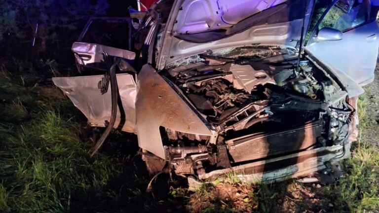 Zniszczony samochód osobowy leży na poboczu drogi, otoczony trawą, z otwartą maską i uszkodzoną przednią częścią.