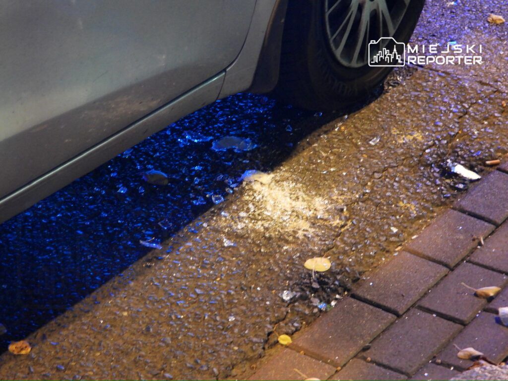Srebrny samochód zaparkowany obok chodnika, obok którego leży rozbita butelka i resztki szkła na mokrej nawierzchni.