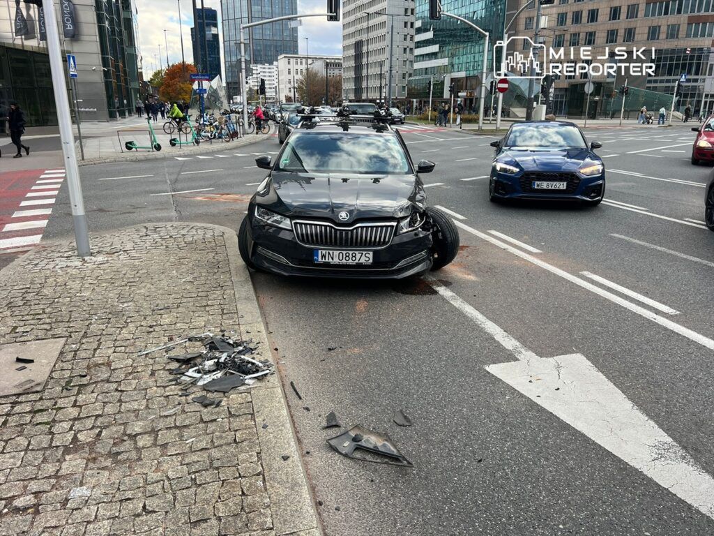 Czarny samochód osobowy z uszkodzoną przednią częścią stoi na jezdni, obok leżą fragmenty karoserii.