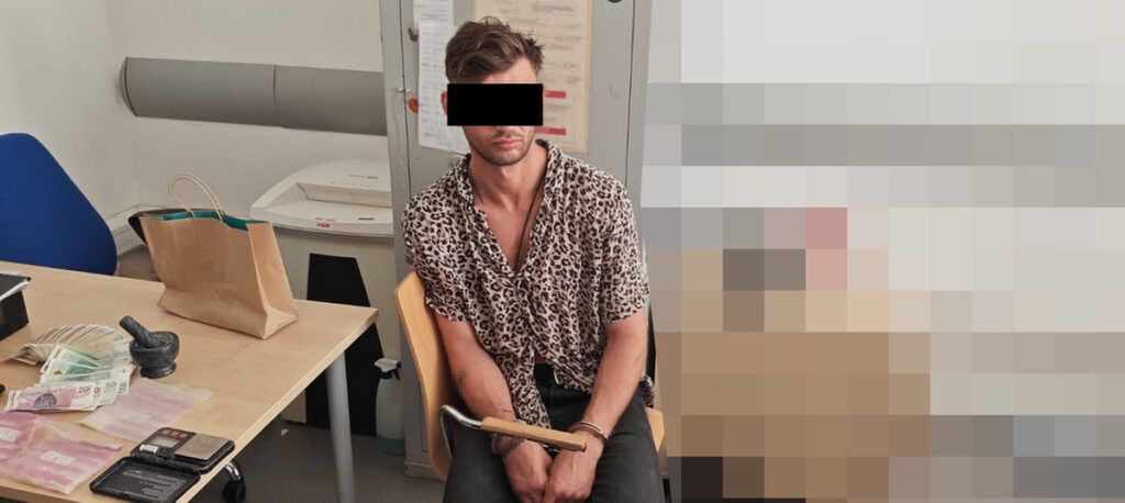 Mężczyzna w koszuli w panterkę siedzi na krześle w pomieszczeniu biurowym, z rękami związanymi metalowym łańcuchem. Na stole leżą pieniądze i przedmioty osobiste.