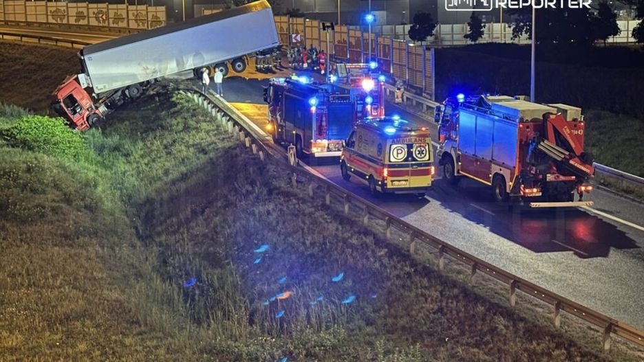 Czerwony ciężarowy pojazd w niebezpiecznej pozycji na poboczu, otoczony przez służby ratunkowe i straż pożarną w nocy.