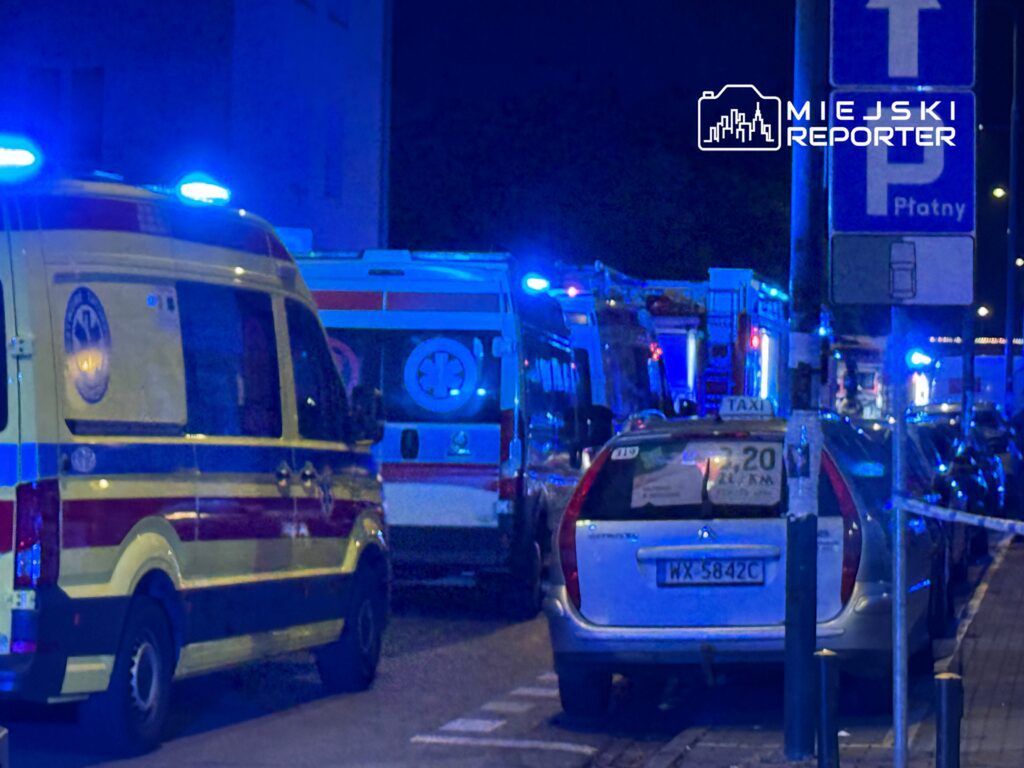 Karetki pogotowia oraz straż pożarna zaparkowane na ulicy, oświetlone niebieskimi sygnałami w nocy.