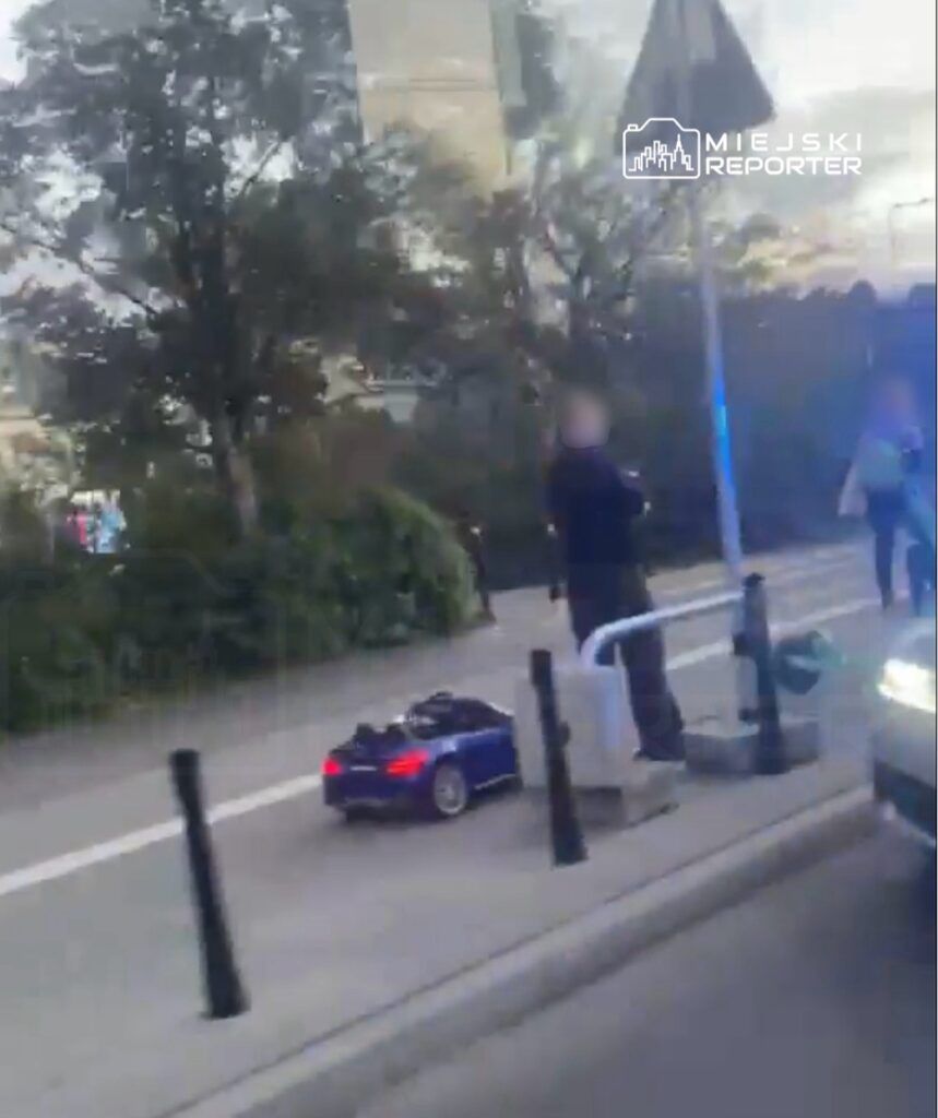 Mężczyzna w czarnym ubraniu obserwujący mały, niebieski samochodzik na chodniku w miejskim otoczeniu.