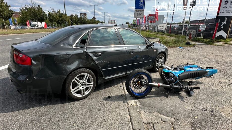 Czarny samochód osobowy z uszkodzeniami na karoserii stoi obok przewróconego motocykla w miejskim otoczeniu.