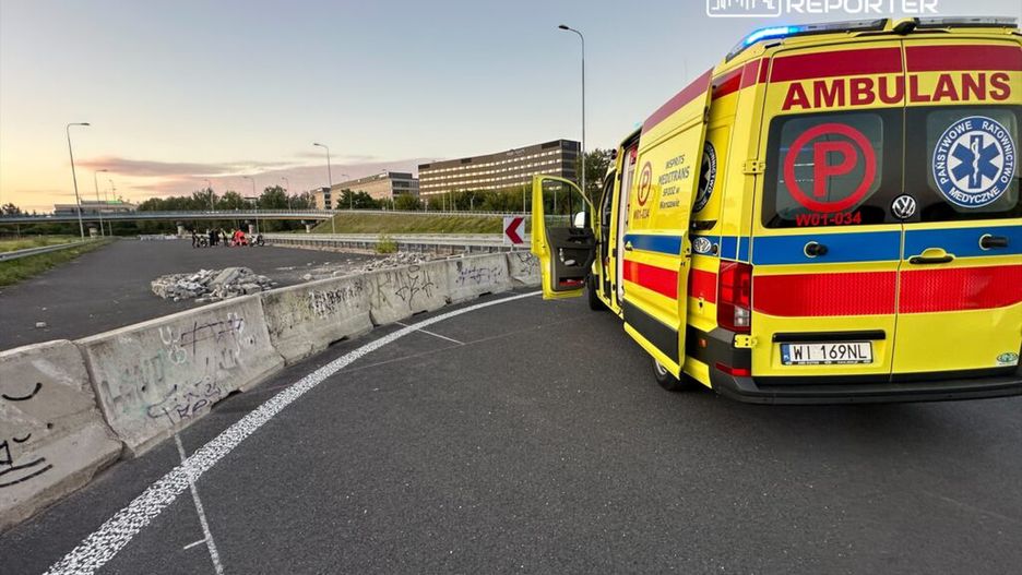 Zespół ratownictwa medycznego przy ambulansie na drodze, obok grupy ludzi i barier betonowych.