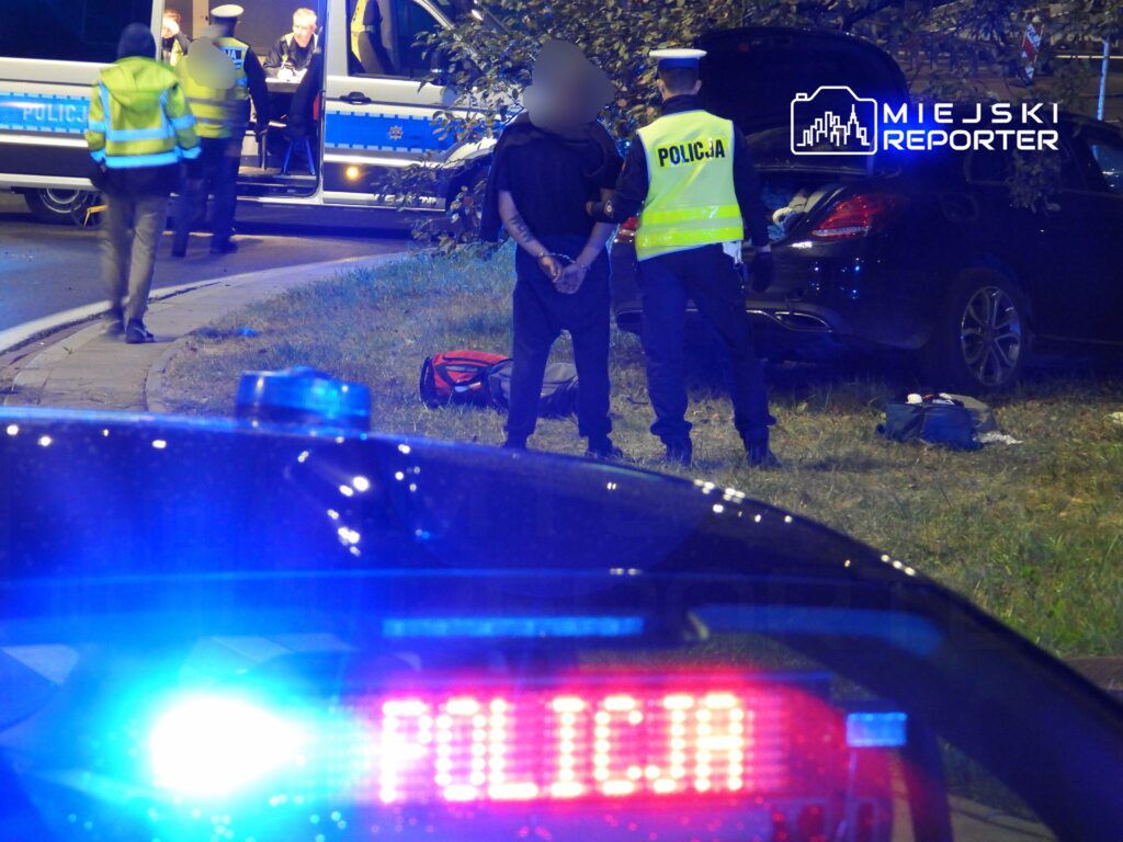 Mężczyzna w kajdankach stoi przed policjantem w kamizelce, obok policyjnego samochodu na poboczu drogi.