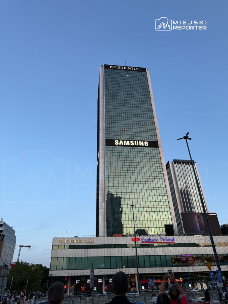 Nowoczesny wieżowiec z przeszkloną fasadą, z logo "Samsung" i "Presidential" na górze, otoczony ludźmi na ulicy.