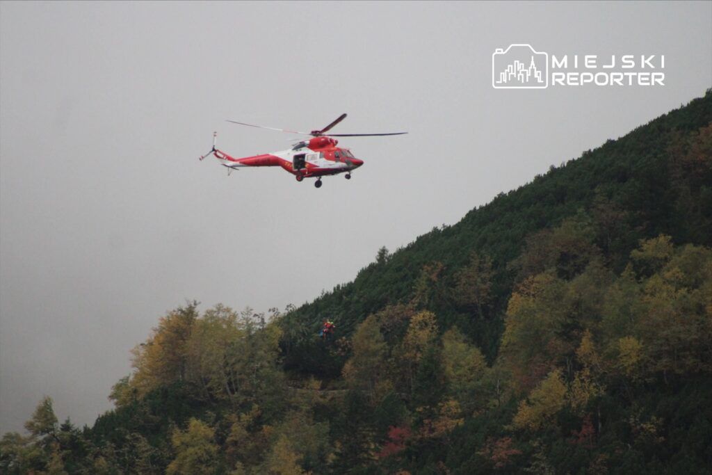 Czerwony helikopter ratunkowy unosi się nad górskim zboczem, z ratownikiem opuszczającym linę.
