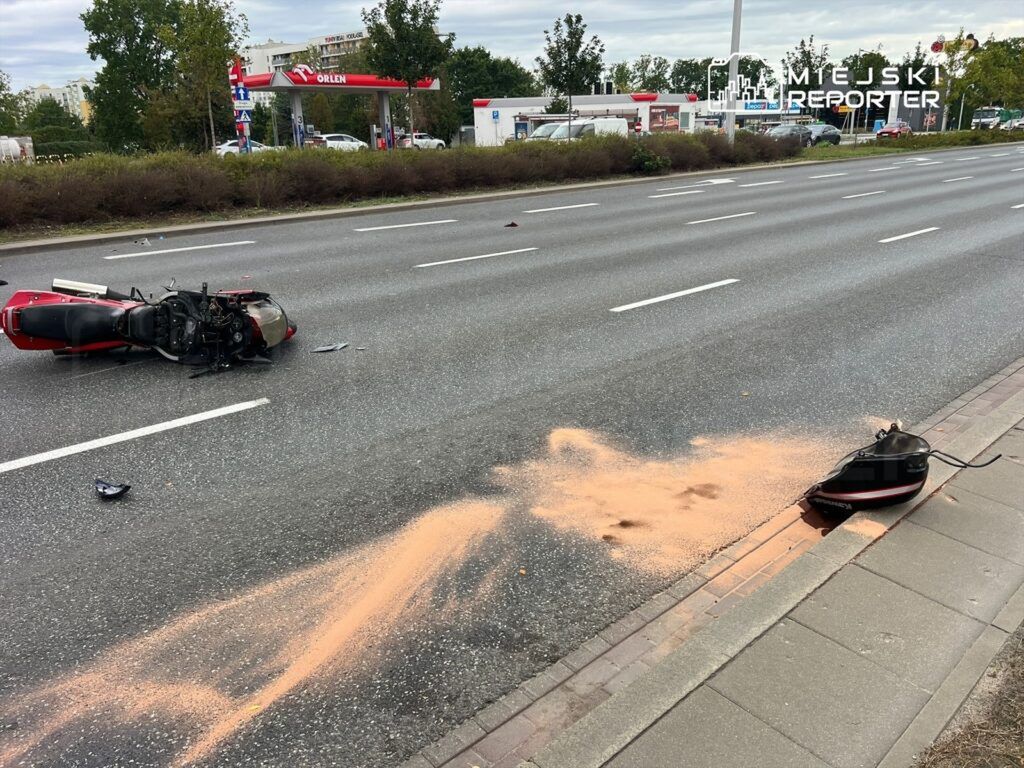 Przewrócony motocykl leży na jezdni, obok rozlana substancja w kolorze pomarańczowym, w tle stacja benzynowa.