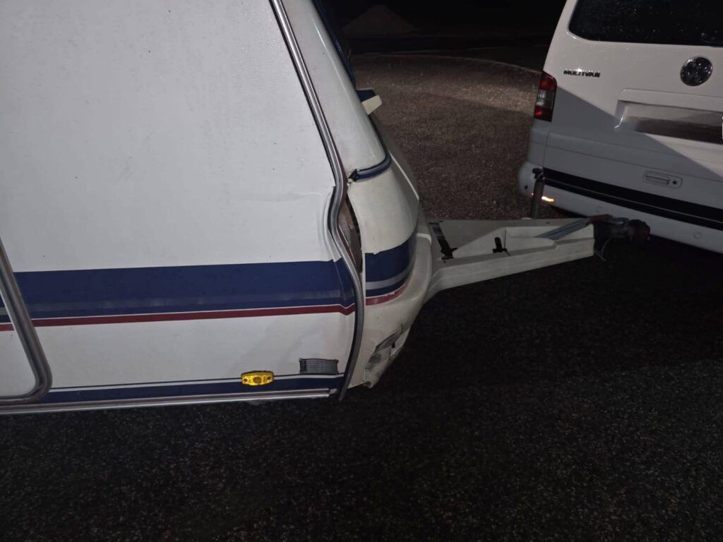 Uszkodzona przyczepa kempingowa z widoczną rysą na boku, przymocowana do samochodu osobowego na nocnym parkingu.