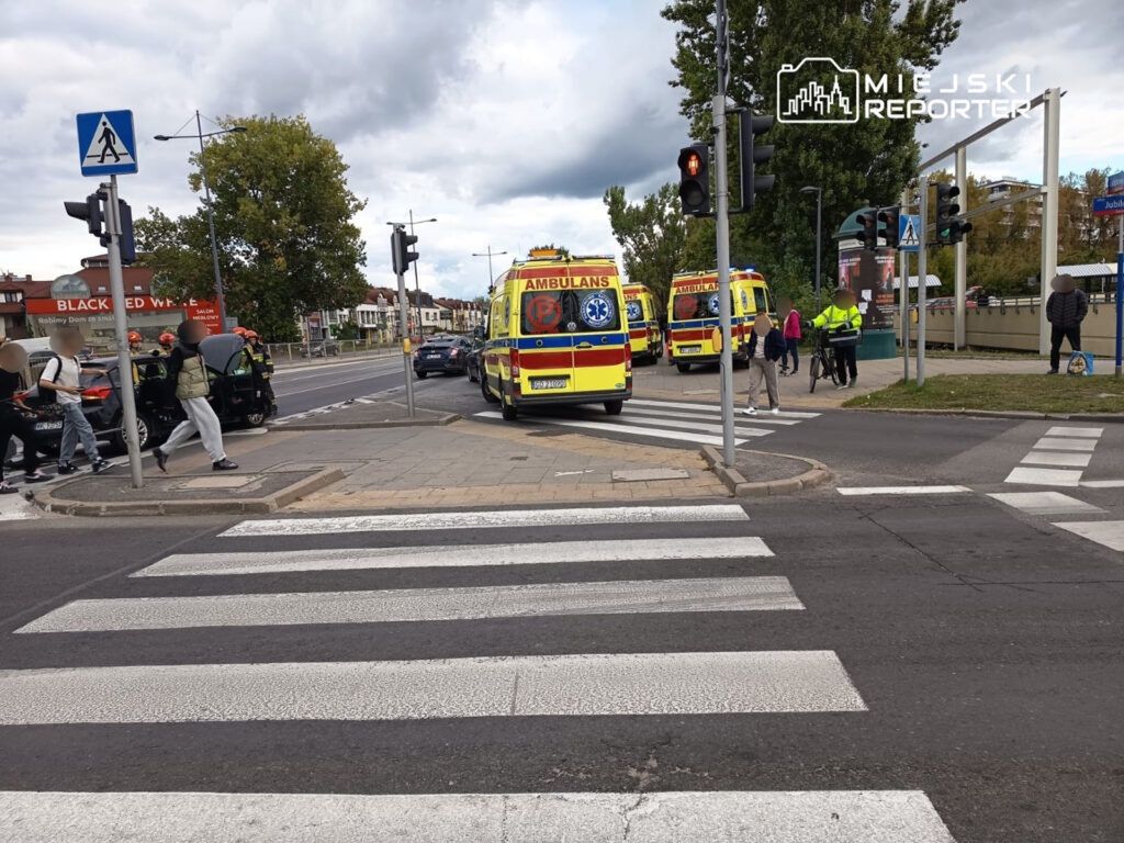 Na przejściu dla pieszych grupa osób przechodzi obok zaparkowanej karetki pogotowia oraz strażaków w hełmach.