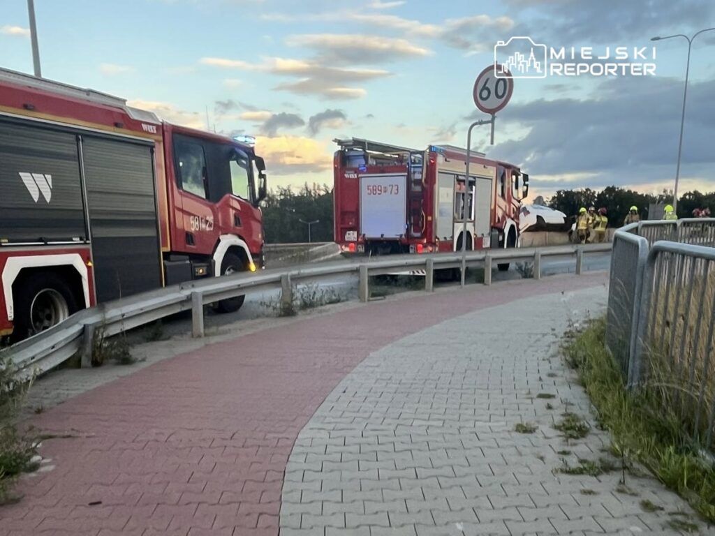 Czerwone wozy strażackie zatrzymane przy drodze, z ratownikami pracującymi nad usunięciem wraku samochodu.
