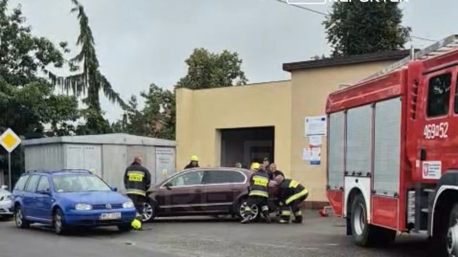 Grupa strażaków w żółtych hełmach pomaga przy usuwaniu pojazdu z wąskiego miejsca obok budynku.