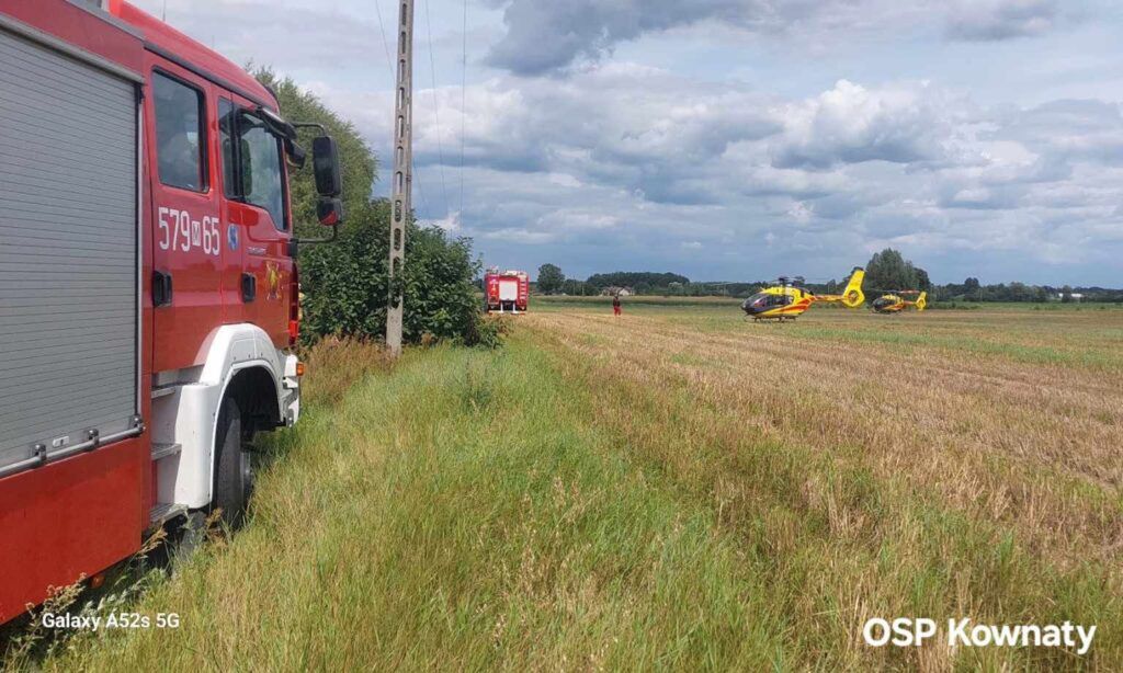 Czerwony wóz strażacki stoi na łące obok dwóch helikopterów ratunkowych, a w tle widoczni ratownicy.