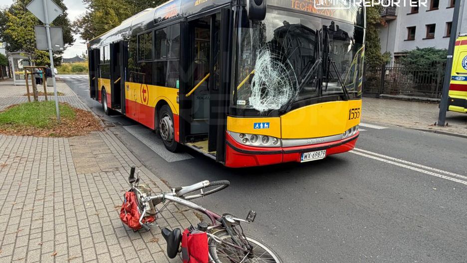 Autobus z uszkodzoną szybą stoi na przystanku, obok leży przewrócony rower z czerwoną torbą.
