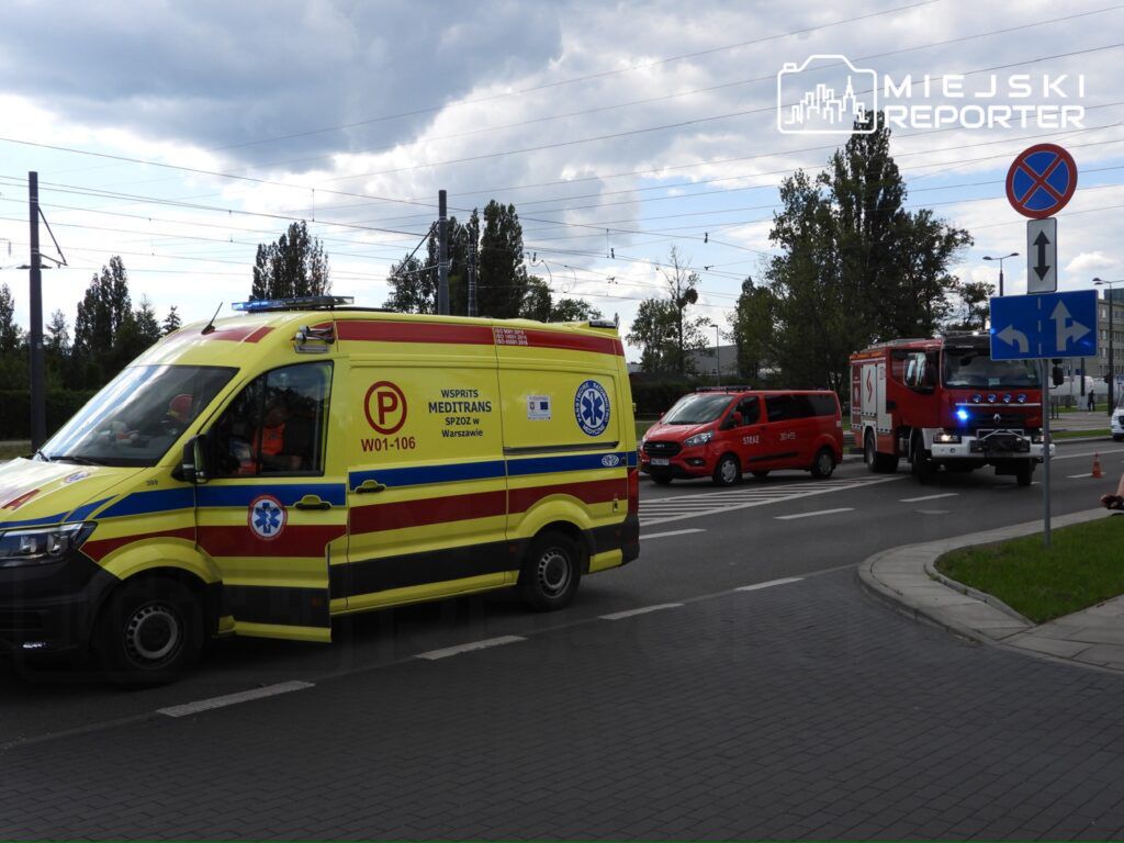 Karetka pogotowia z włączonymi sygnałami przejeżdża obok straży pożarnej i samochodu dostawczego na skrzyżowaniu.
